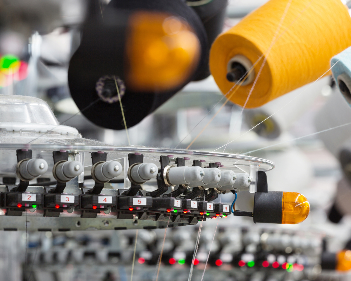 It's a close up shot of a machine that potential separates strings of yarn. It contains numbers from 2 to 9. Yellow yarn and what can be either light grey or very pale light blue yarn are being pulled. An Orange light bulb is attached to the side. 